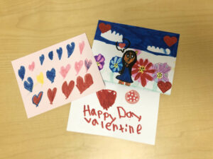 Three valentines cards are displayed on a light wooden background. One card is pink with hand-drawn, multicolored hearts. The second card features a drawing of a girl and large flowers, with a blue sky and white clouds above her. The third card shows a hand-drawn heart with a flower sticker and "Happy Day Valentine" written in red.
