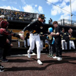 A Missouri baseball player exits the dugout. He is high-fiving kids from the Thompson Center. The sky is blue and there are clouds.