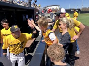 A mom holds her son as they face the Mizzou Tigers baseball dugout. A Tigers player is high-fiving the boy.