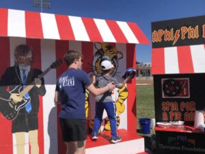 A man and a young boy stand in front of a red and white striped carnival booth. The boy is holding a pie and is going to throw it at the face of someone behind a Truman the Tiger cut-out.