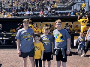 Three boys and one girl are smiling and looking at the camera. They are standing on the baseball field in front of the Mizzou dugout. 