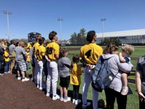 Children stand next to Mizzou Baseball players as they look toward the American flag during the National Anthem. They are standing on the baseball field and are facing away from the camera. It is a sunny day with a blue sky.