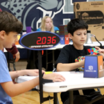 Two boys sit inside at a table. One is solving a Rubik's cube and the other is watching him, serving as a judge. There is a large stopwatch attached to the table that shows the cuber's time.