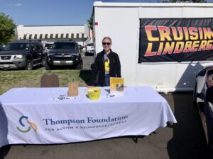 A white woman stands in the middle of the picture. She is wearing sunglasses, a black jacket, and is smiling. She is standing outside behind a white tablecloth that has the Thompson Foundation logo on the front.