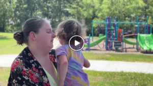 A woman holds her daughter at a playground. There is a play button in the middle of the picture.