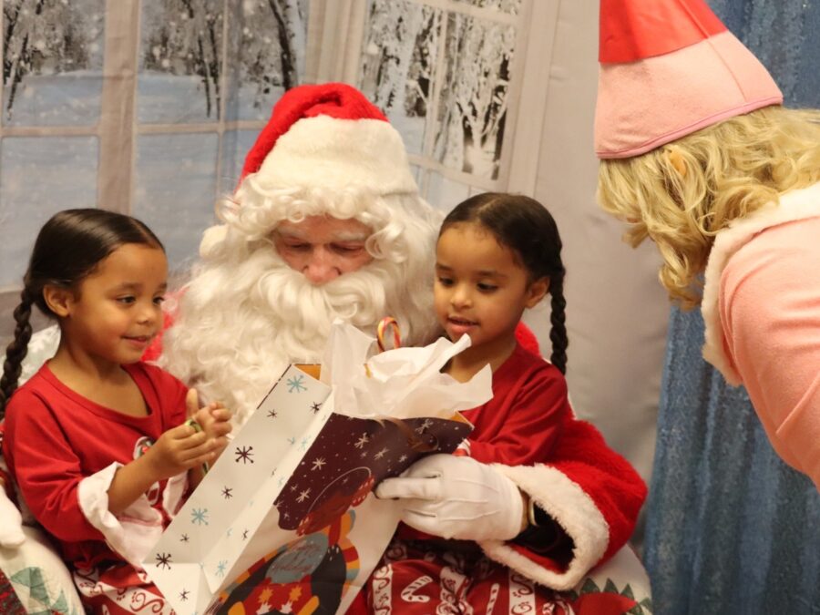 Two girls with braided brown hair sit on Santa's lap. He is holding a gift and they are smiling. Santa's elf looks on.
