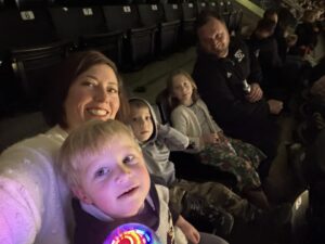 A family with a mom, dad, and three kids takes a selfie in an arena.